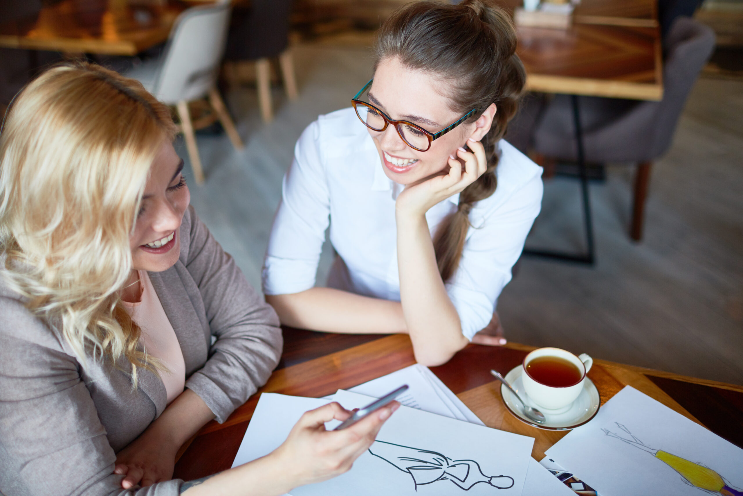 Joyful young fashion designers having fun while discussing joint project at cozy small coffeehouse, pretty blond-haired woman using smartphone